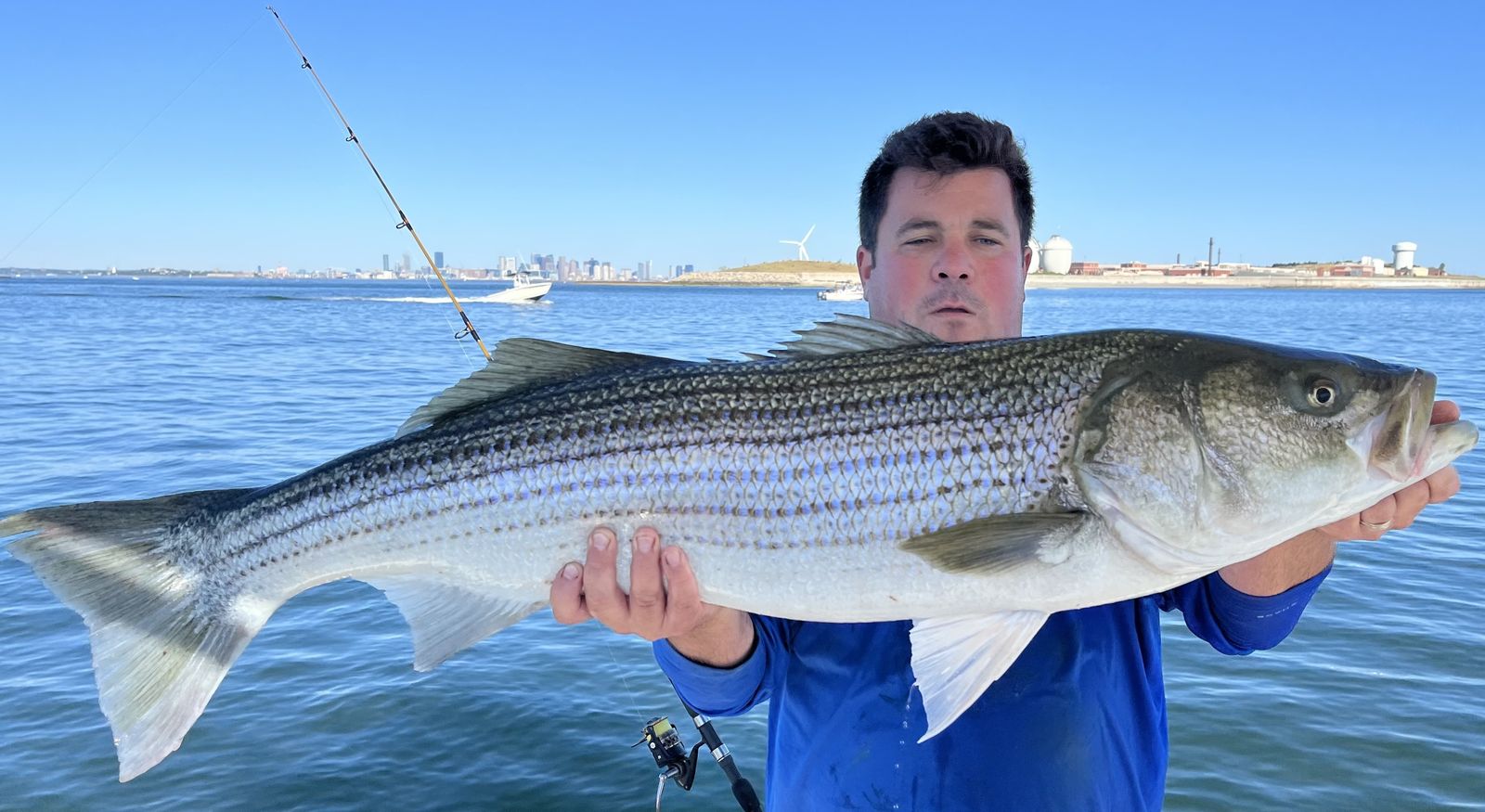A trophy striped bass held up with the Boston skyline across the water