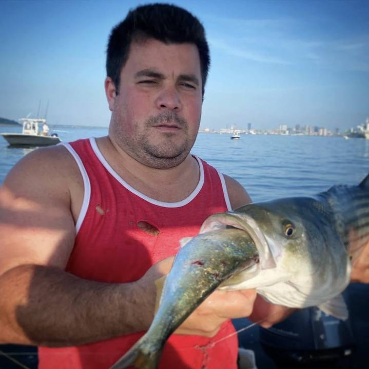 A bluefish held up close with the shoreline behind